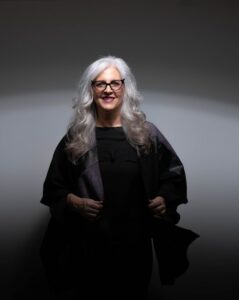 A woman with silver hair and black glasses stands in a dark room